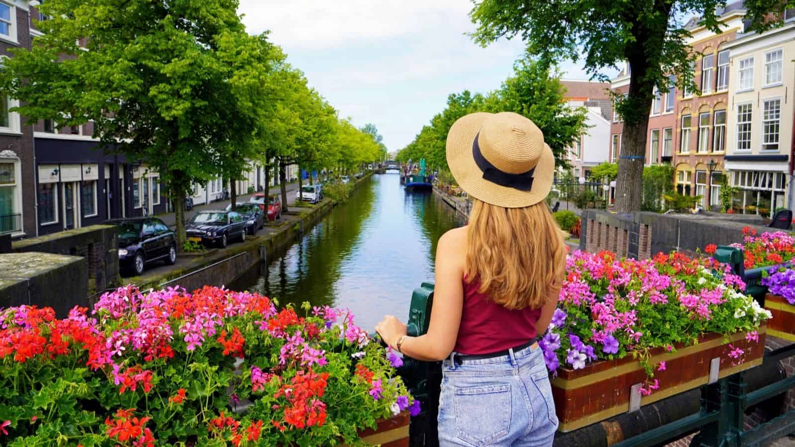 Woman in a hat stands on a bridge with flowers, looking at a canal lined with trees and buildings.