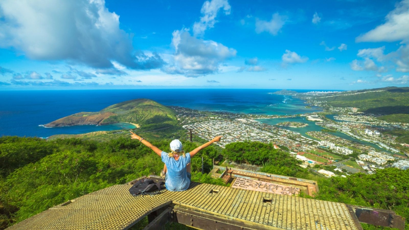 Person sitting on a platform with arms raised, overlooking a coastal city and ocean under a blue sky.