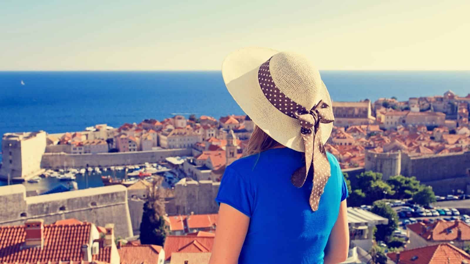 Woman in a sunhat overlooks a coastal town with terracotta roofs and blue sea under a clear sky.