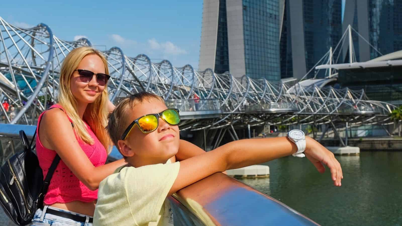 A woman and a boy in sunglasses lean on a bridge with modern city buildings in the background.