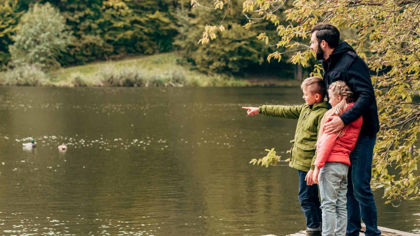 Man with two children stands by a lake. One child points across the water; trees in background.