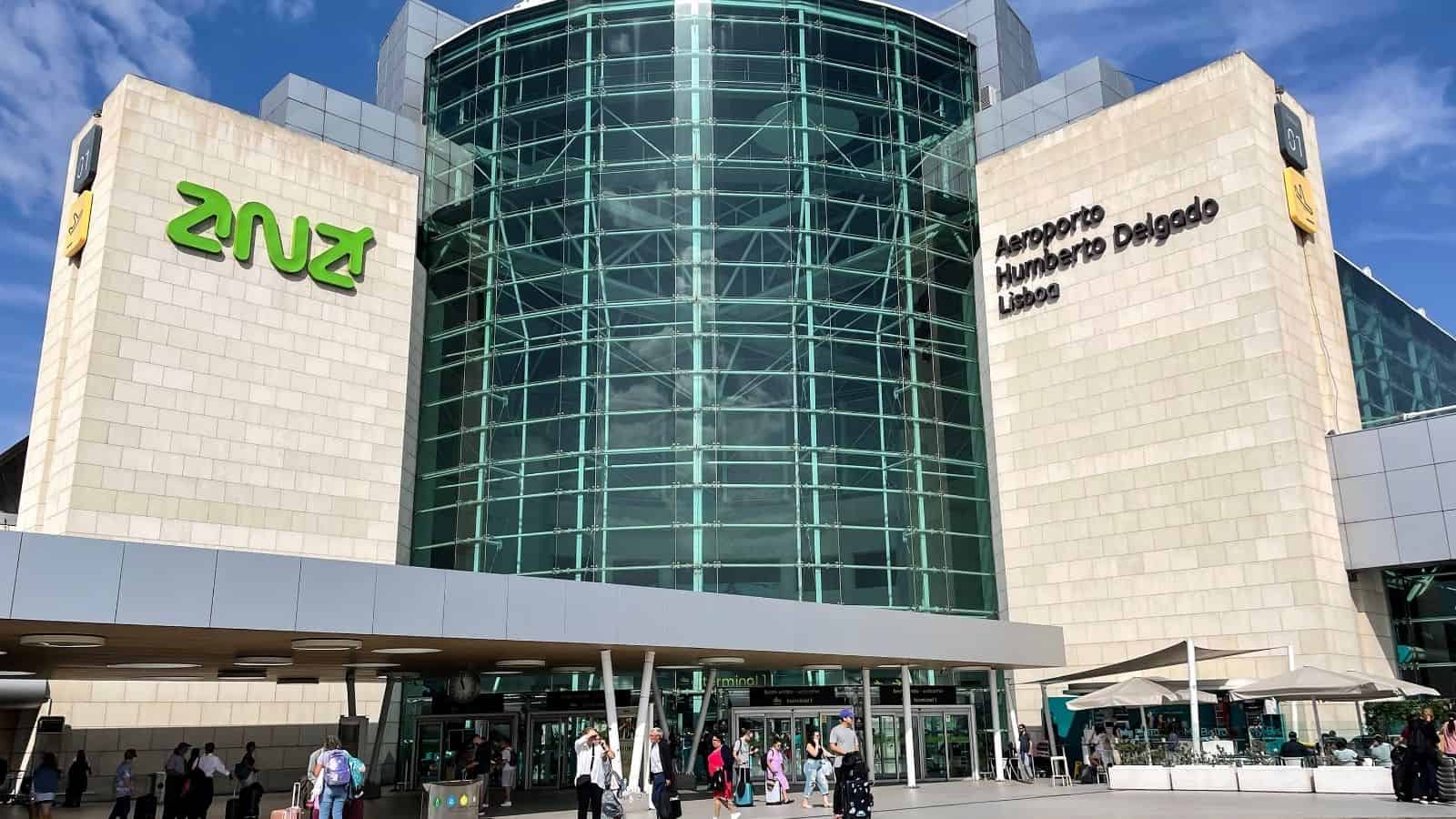 Glass entrance of Aeroporto Humberto Delgado Lisboa with people walking and outdoor seating visible.