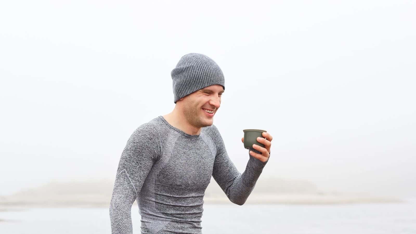 Man in a gray beanie and shirt smiles while holding a cup outdoors on a foggy day, ready for unpredictable weather with his travel essentials.