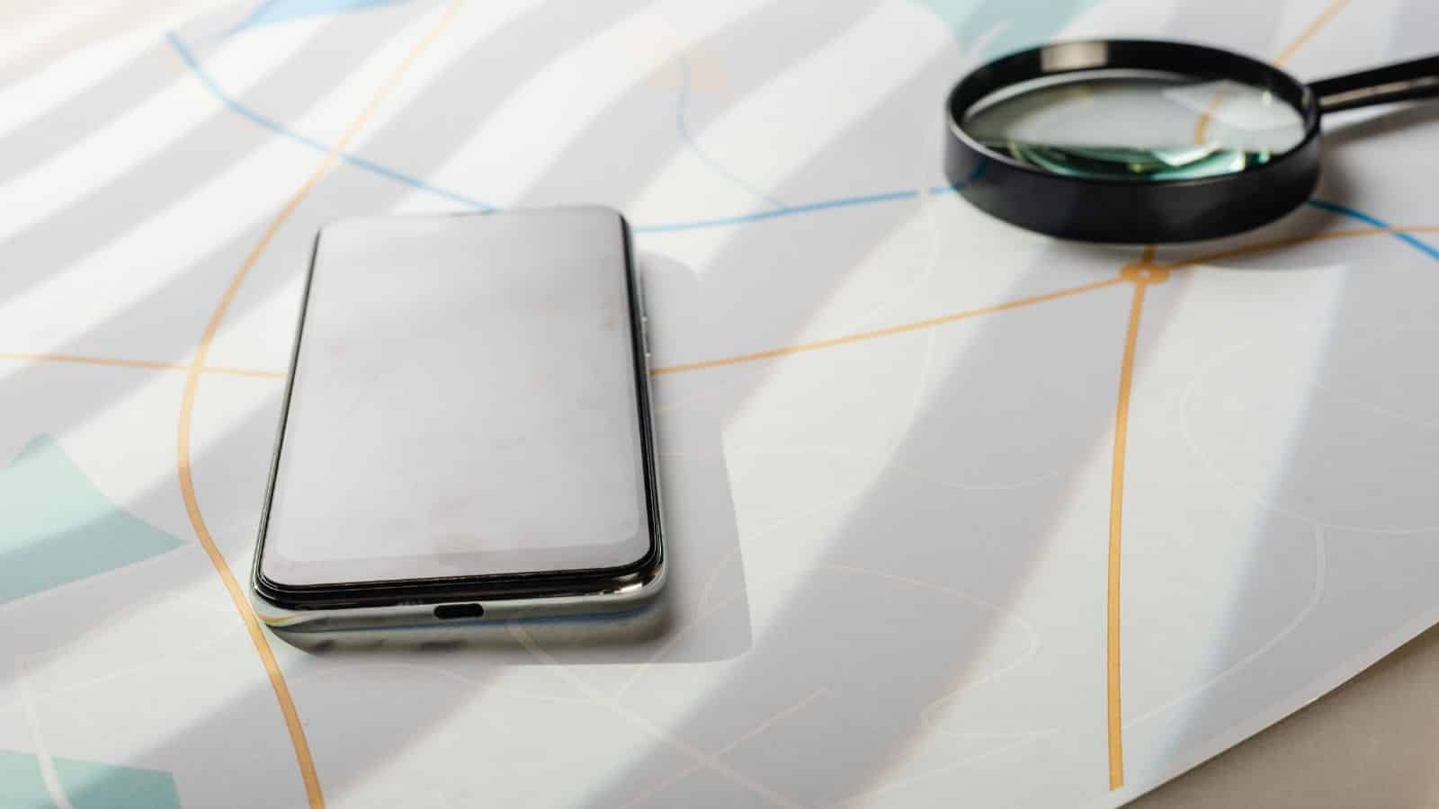 A smartphone and a magnifying glass rest on a paper map with colored lines, in sunlight—true travel essentials for navigating new places, especially when facing unpredictable weather.