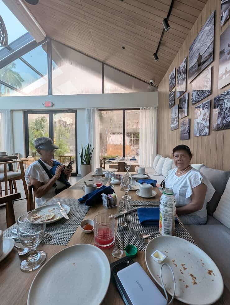 Two women sit at a restaurant table with finished meals and drinks, sunlight streaming through large windows, sharing stories from their unforgettable Belize reef and ruins trip.