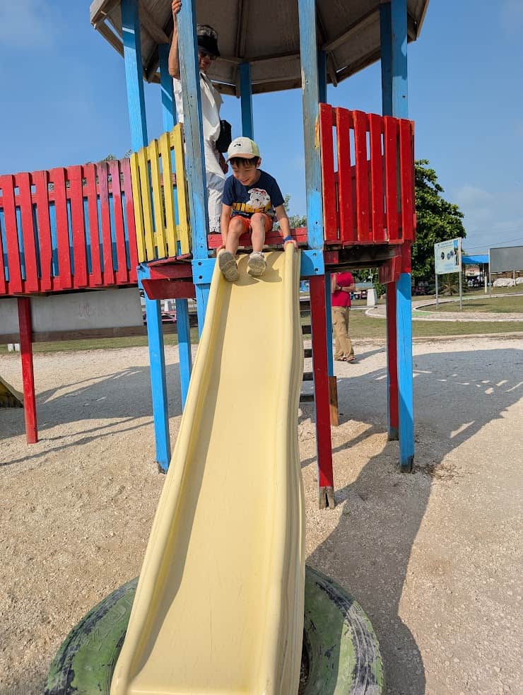 Child wearing a hat sits at the top of a yellow slide on a colorful playground, while an adult stands behind&mdash;ready for adventure just like on a Belize reef and ruins trip.