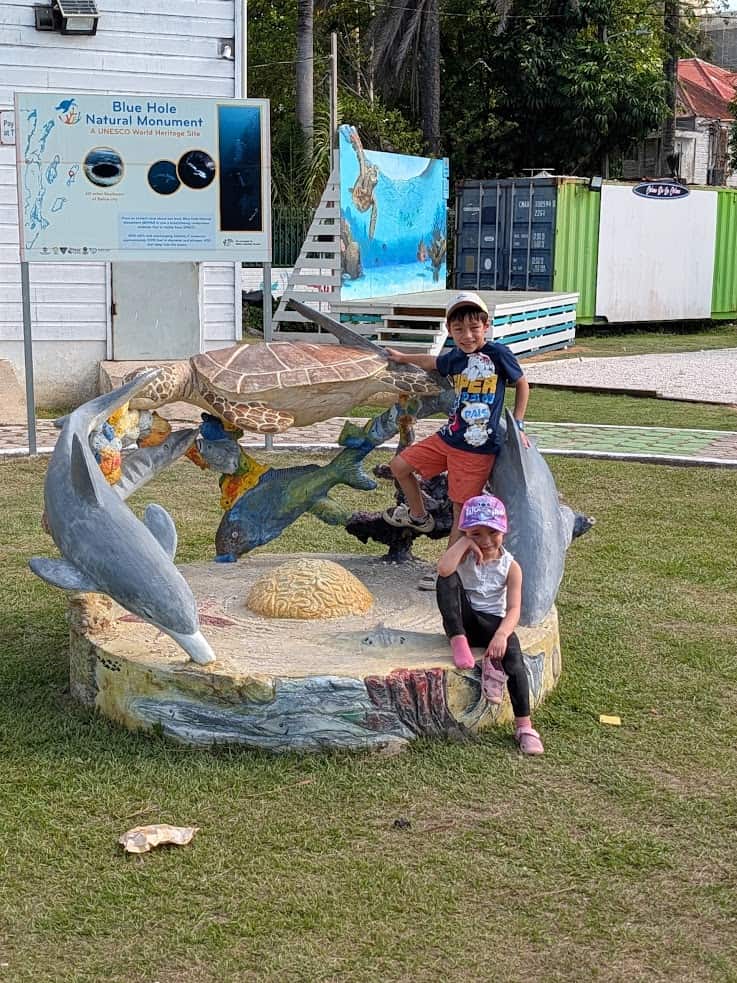 Two kids pose on a turtle and dolphin statue in front of a Blue Hole Natural Monument sign outdoors, capturing the spirit of adventure on their unforgettable Belize reef and ruins trip.