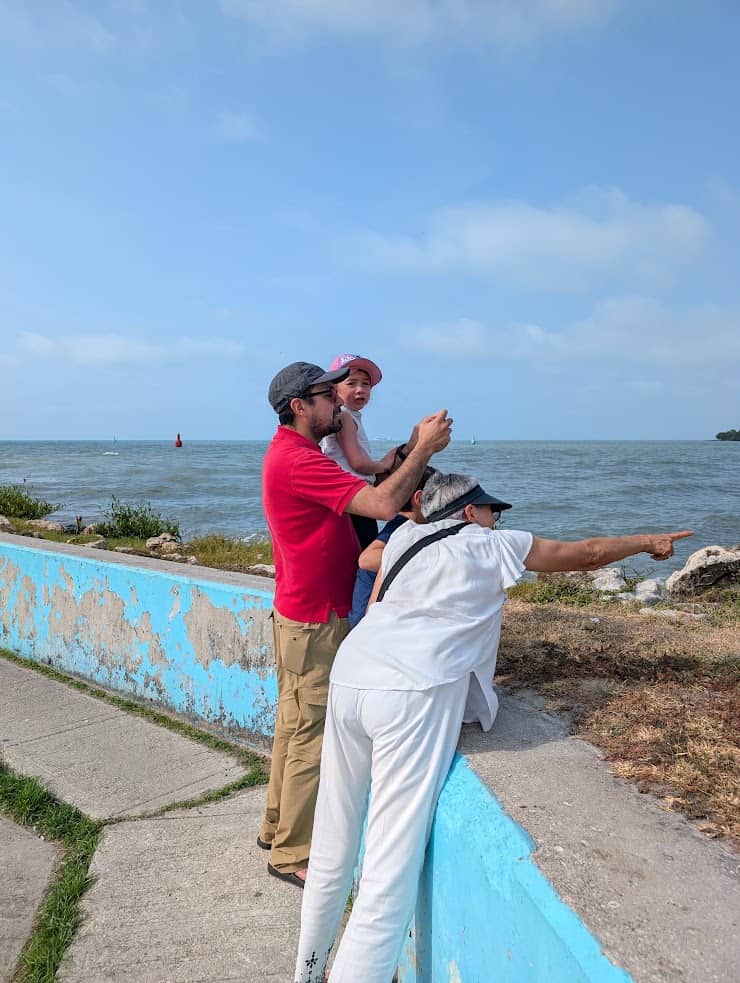 Three people by a seaside wall; a man holds a child while another man in white points toward the ocean, perhaps sharing stories from their recent Belize reef and ruins trip.