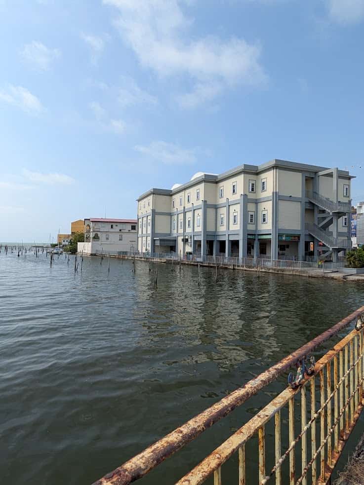 A light blue building on stilts stands over calm water, with a rusty railing in the foreground&mdash;a tranquil scene reminiscent of views from a Belize reef and ruins trip.
