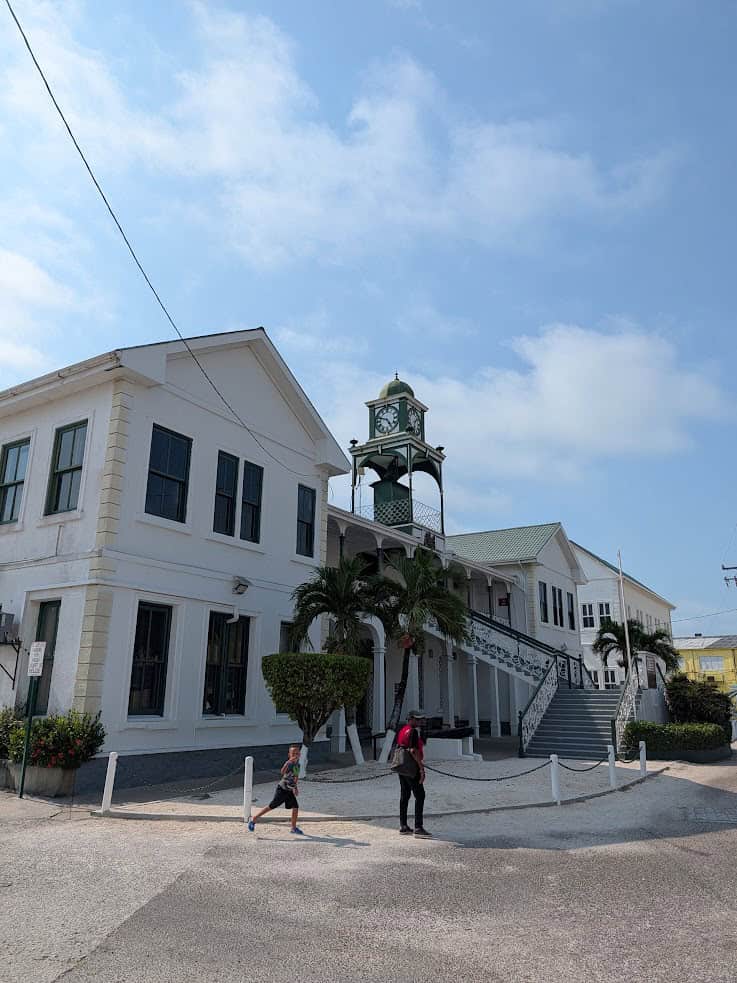 White colonial building with a clock tower, palm trees, and two people walking in front on a sunny day&mdash;a classic scene you might enjoy before or after your Belize reef and ruins trip.