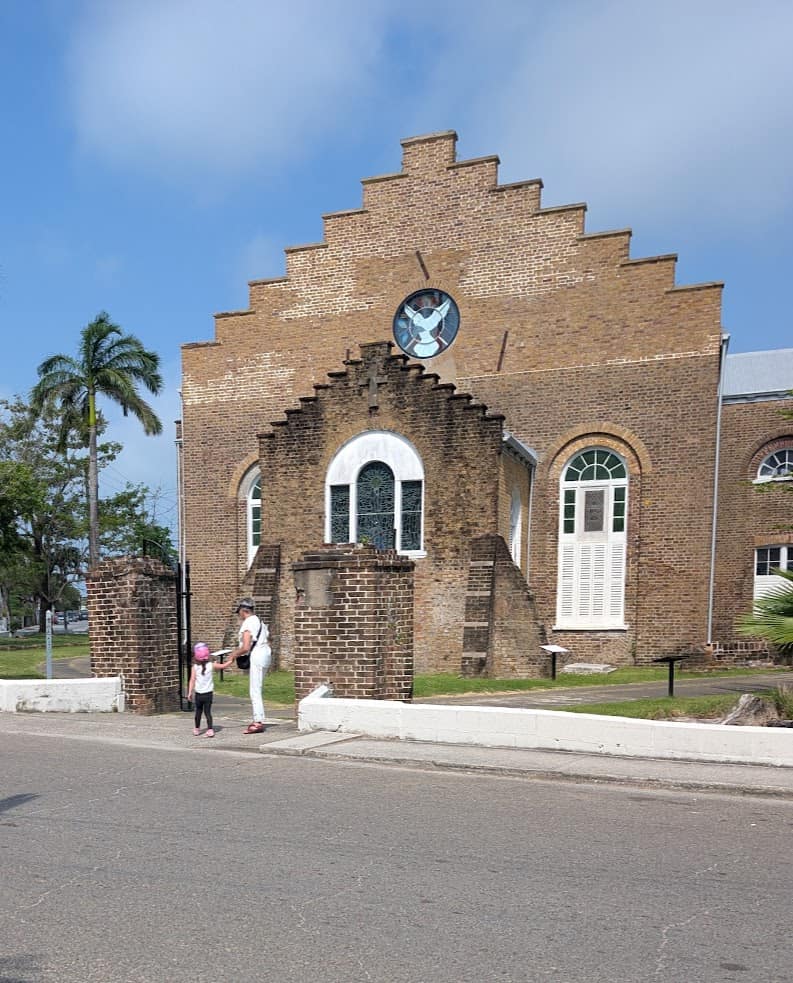 Two people stand outside a brown brick church with arched windows and a circular stained glass window, reminiscing about their recent Belize reef and ruins trip.