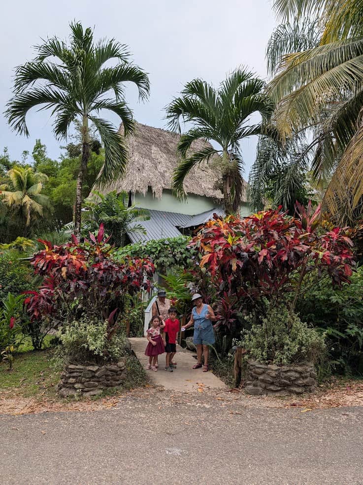 Three people stand on a garden path in front of a house with a thatched roof, surrounded by tropical plants, reminiscent of the lush setting described in The Rainforest Lodge at Sleeping Giant Review.