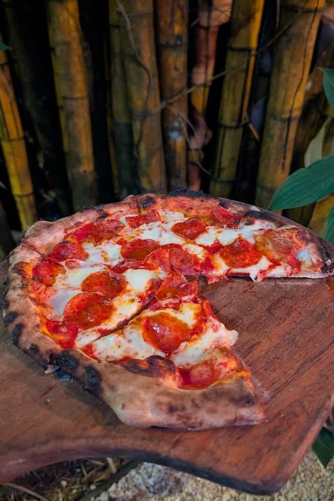 A pepperoni pizza with one slice missing sits on a wooden board, surrounded by plants and bamboo, evoking the relaxed atmosphere of a Belize reef and ruins trip.
