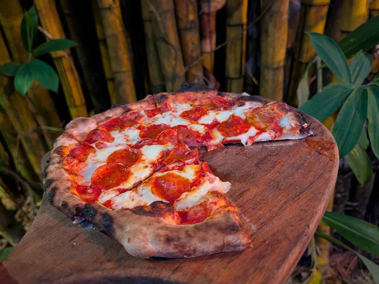 A pepperoni pizza with a missing slice sits on a wooden board in front of bamboo and green plants, creating a scene reminiscent of an outdoor feast at The Rainforest Lodge at Sleeping Giant.