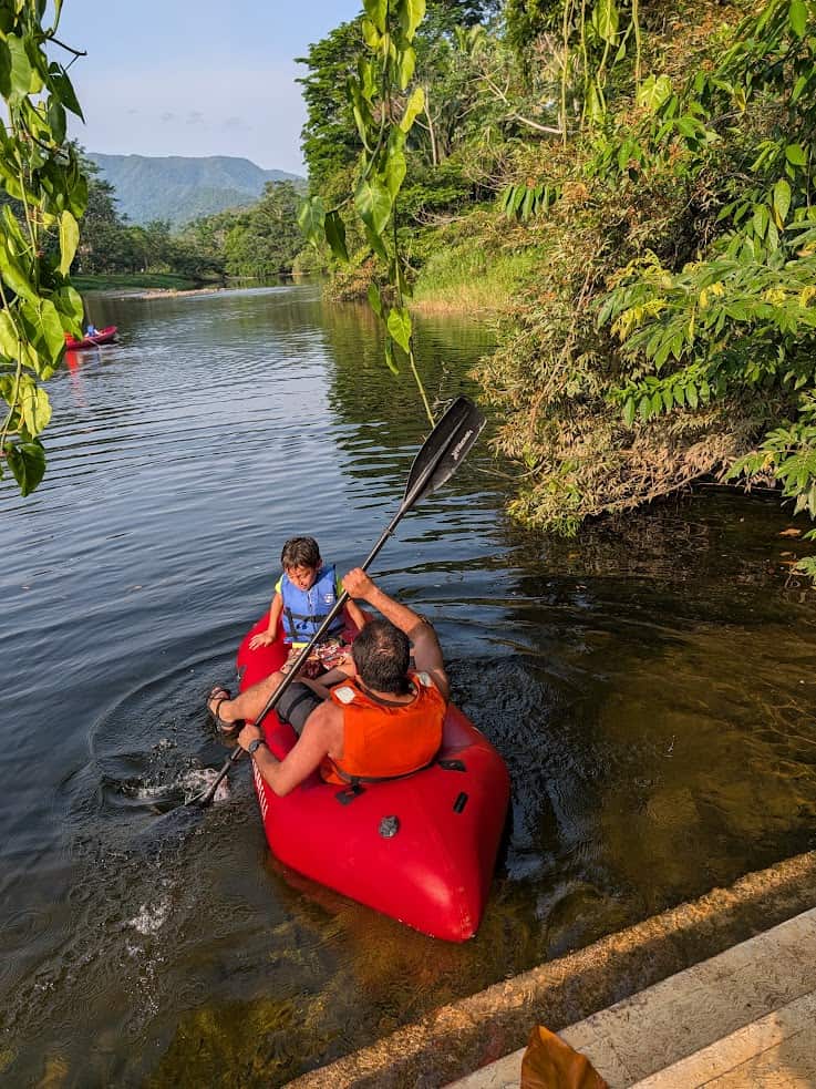 Two people in a red kayak paddle near a leafy riverbank, one adult and one child wearing life jackets—an adventure that could easily be part of The Rainforest Lodge at Sleeping Giant Review, capturing the spirit of exploration and natural beauty found at this unique destination.
