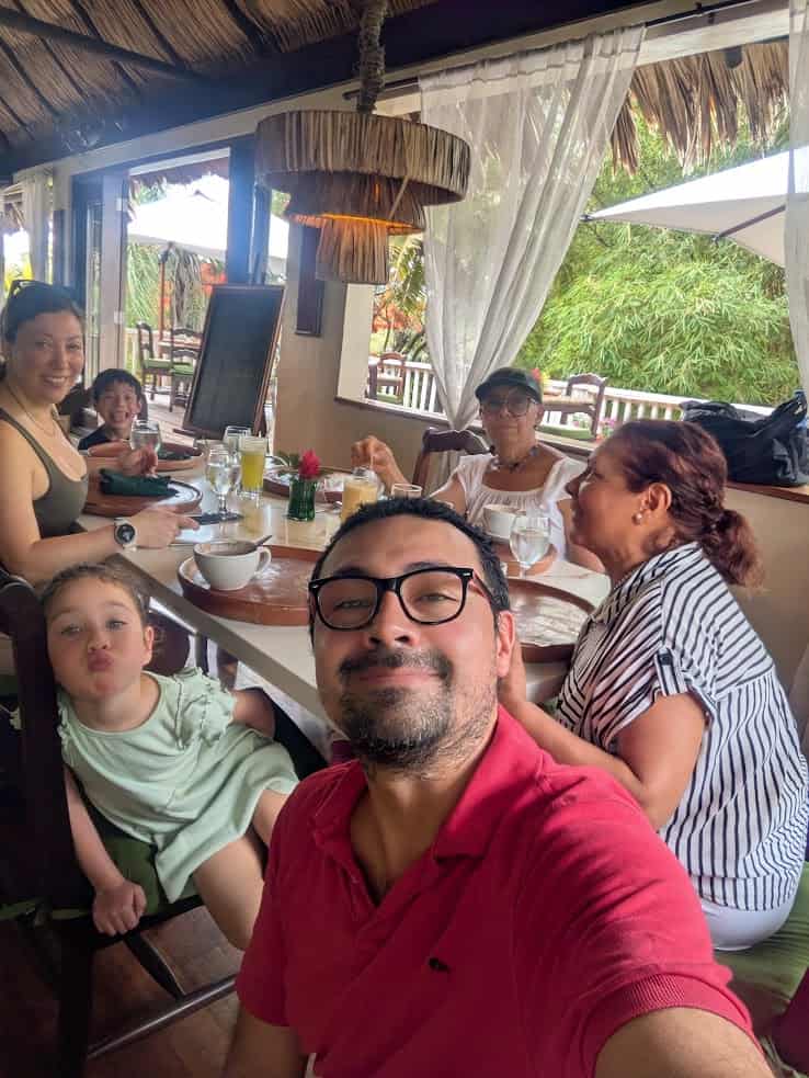 Six people of various ages smiling and sitting around a restaurant table with drinks and food, enjoying a lively meal together reminiscent of the warm atmosphere described in The Rainforest Lodge at Sleeping Giant Review.
