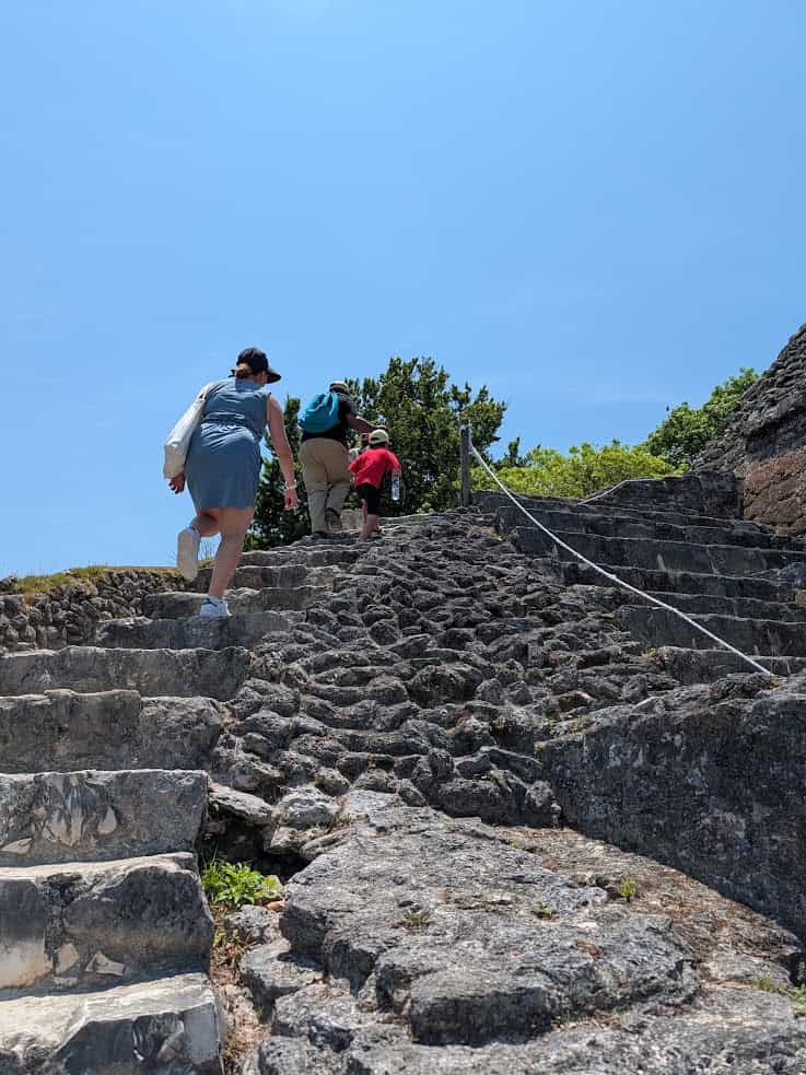 Three people climb uneven stone steps outdoors under a bright blue sky, surrounded by greenery and trees&mdash;an experience reminiscent of a Belize reef and ruins trip.
