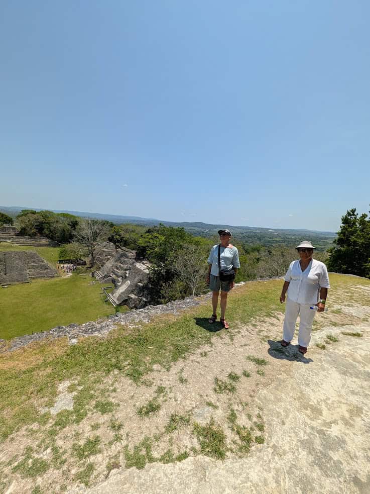 Two people stand on a rocky hill overlooking ancient ruins and green trees under a clear sky, capturing the spirit of a Belize reef and ruins trip.