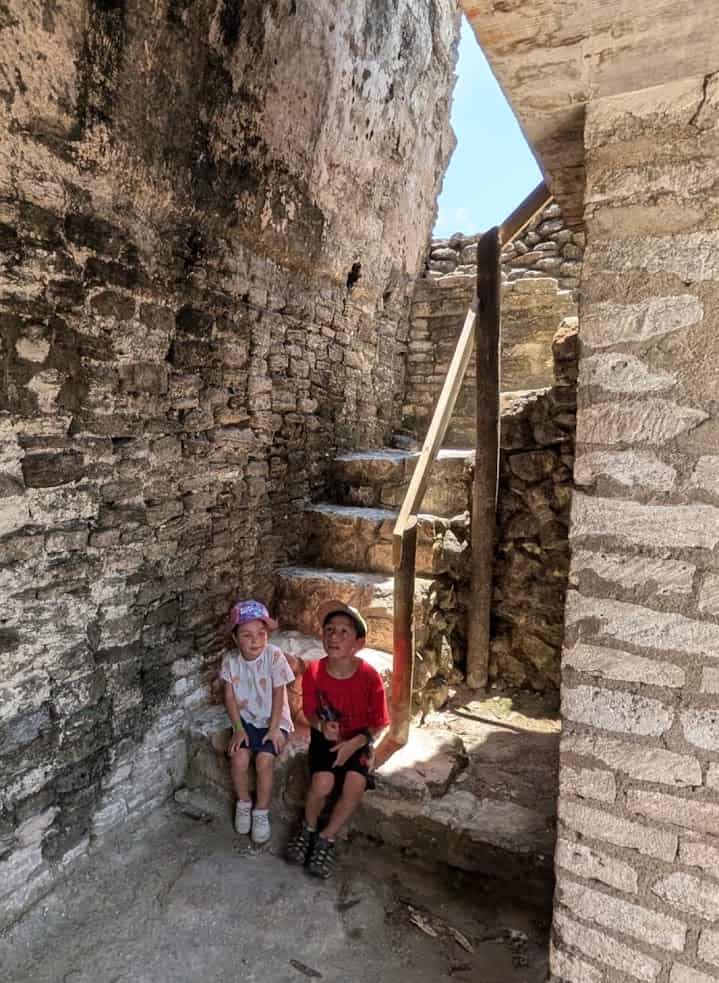 Two kids sit on stone steps in a sunlit, ancient ruin with weathered walls and a wooden handrail, capturing the spirit of adventure found on a Belize reef and ruins trip.