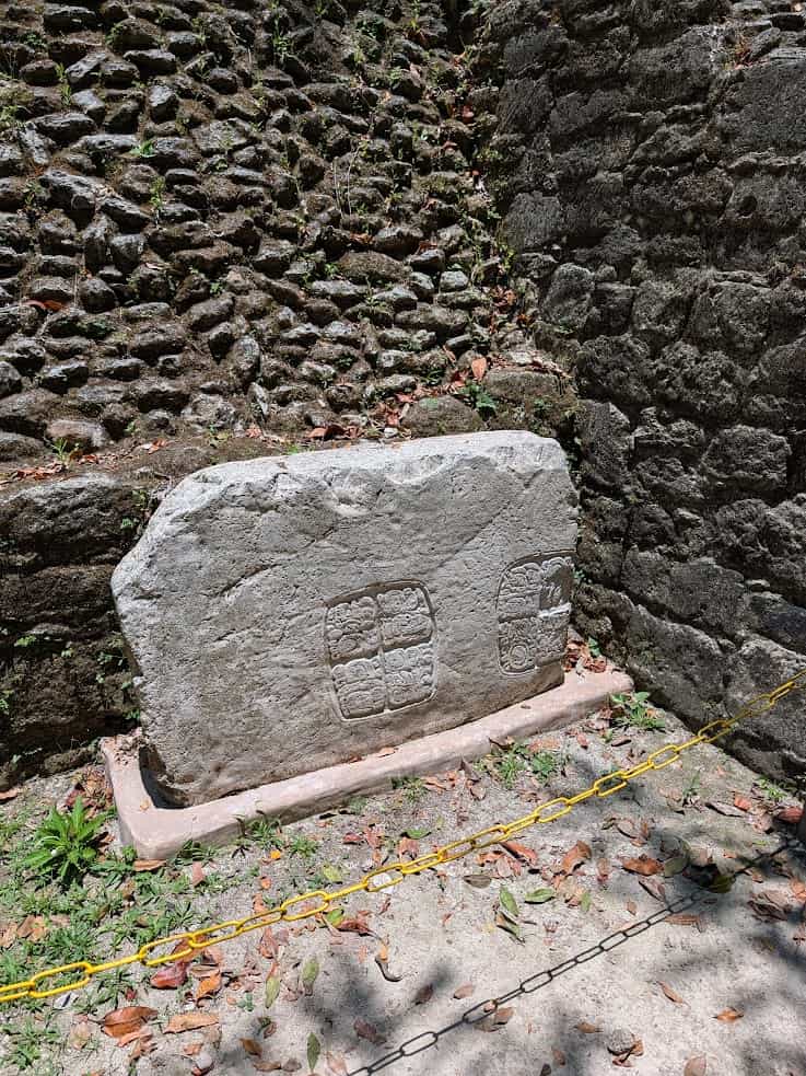 Stone slab with ancient carvings, set against a stone wall and surrounded by yellow caution chains&mdash;a mysterious relic reminiscent of discoveries on a Belize reef and ruins trip.