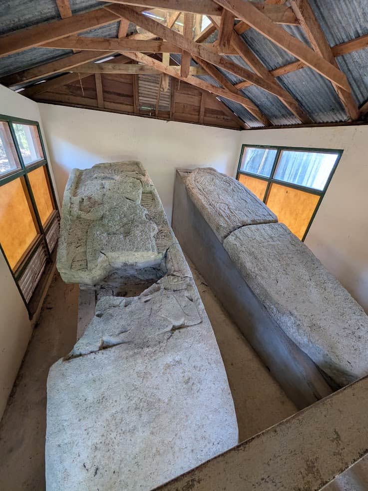 Two large stone sarcophagi rest inside a small building with a corrugated metal roof and windows, offering a glimpse into the ancient history you might encounter on a Belize reef and ruins trip.