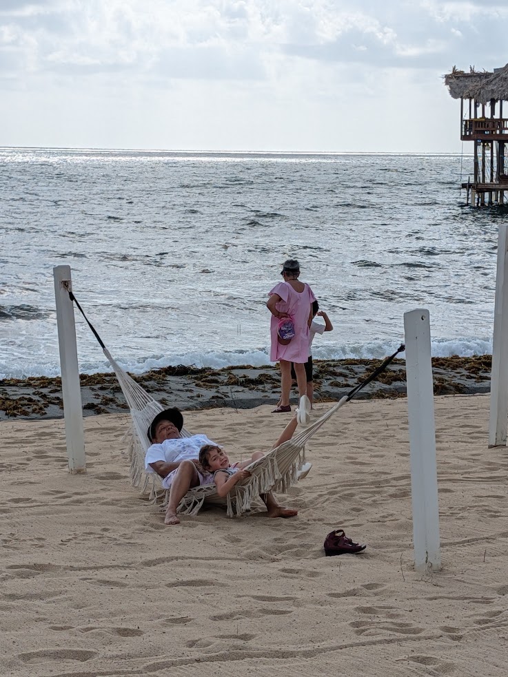 Two people relax on a hammock on a sandy beach; another person stands nearby with the ocean in the background, embodying the tranquil scene described in many a The Lodge at Jaguar Reef review.