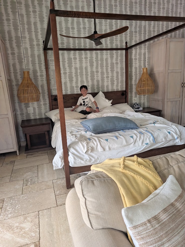 A child sits on a canopy bed in a cozy bedroom with rattan lamps and light-colored furniture, evoking the relaxed comfort found in a glowing The Lodge at Jaguar Reef review.