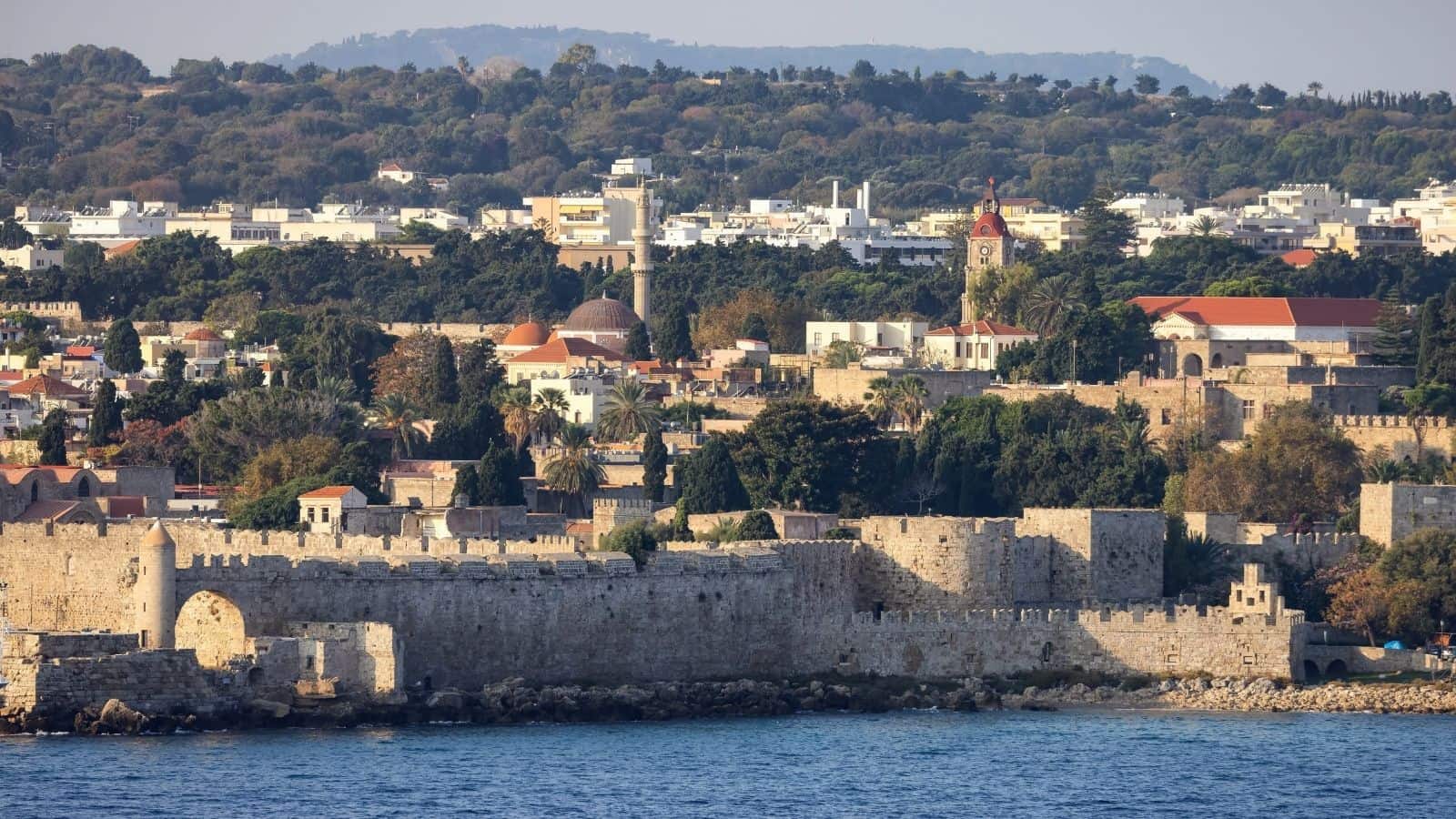 A stone fortress with arched gates by the sea, historic buildings and trees in the background.