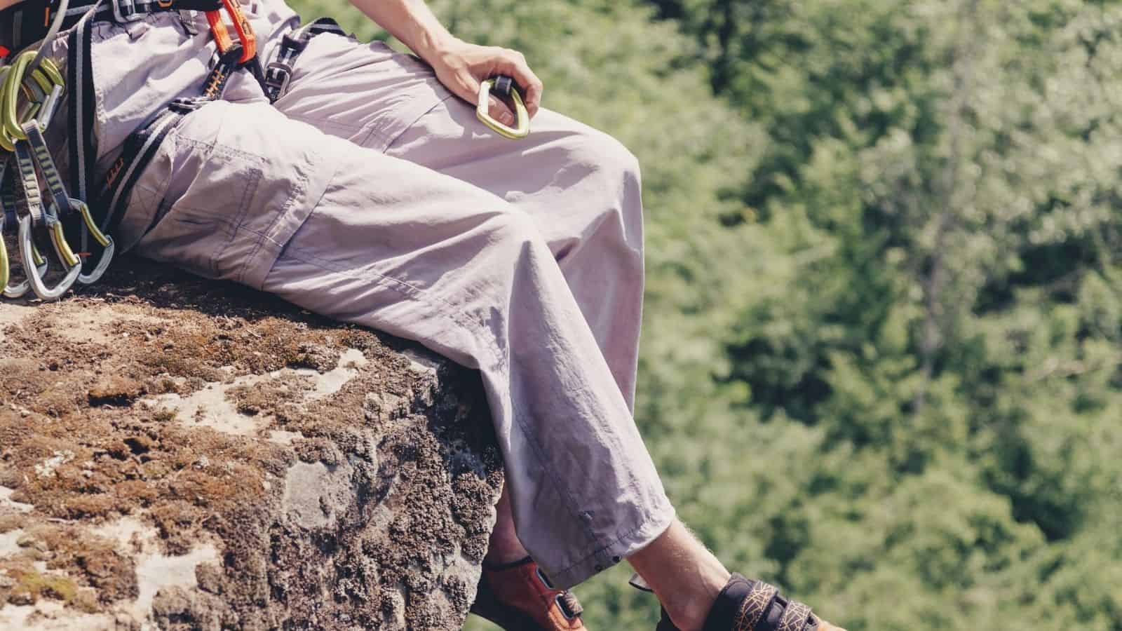 Person in climbing gear sitting on a rocky ledge, holding a carabiner—surrounded by trees in the background—pauses to reflect on their hiking journey and the importance of packing travel essentials, especially when facing unpredictable weather.