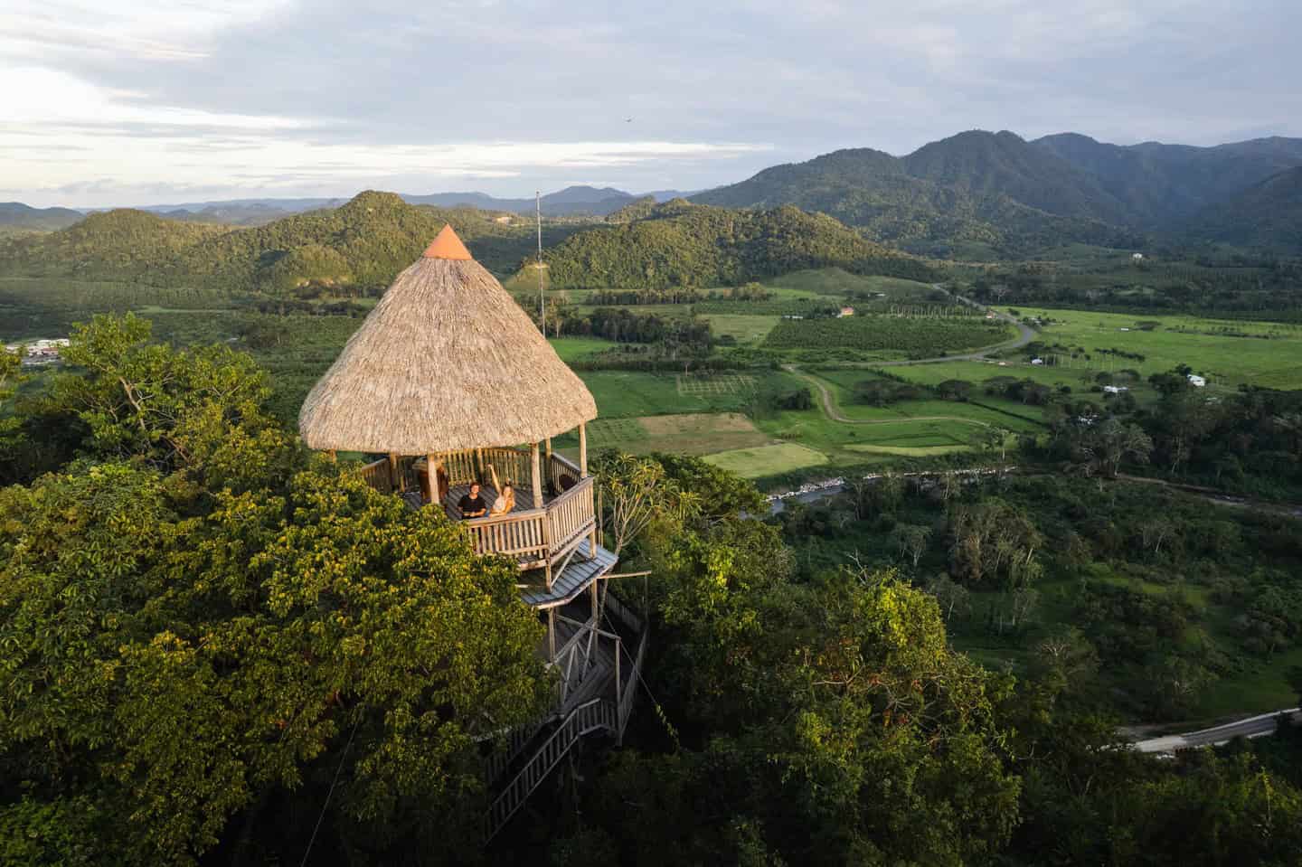 Thatched-roof treehouse overlooks lush green valley and distant mountains under a partly cloudy sky, evoking the serene ambiance described in The Rainforest Lodge at Sleeping Giant Review.