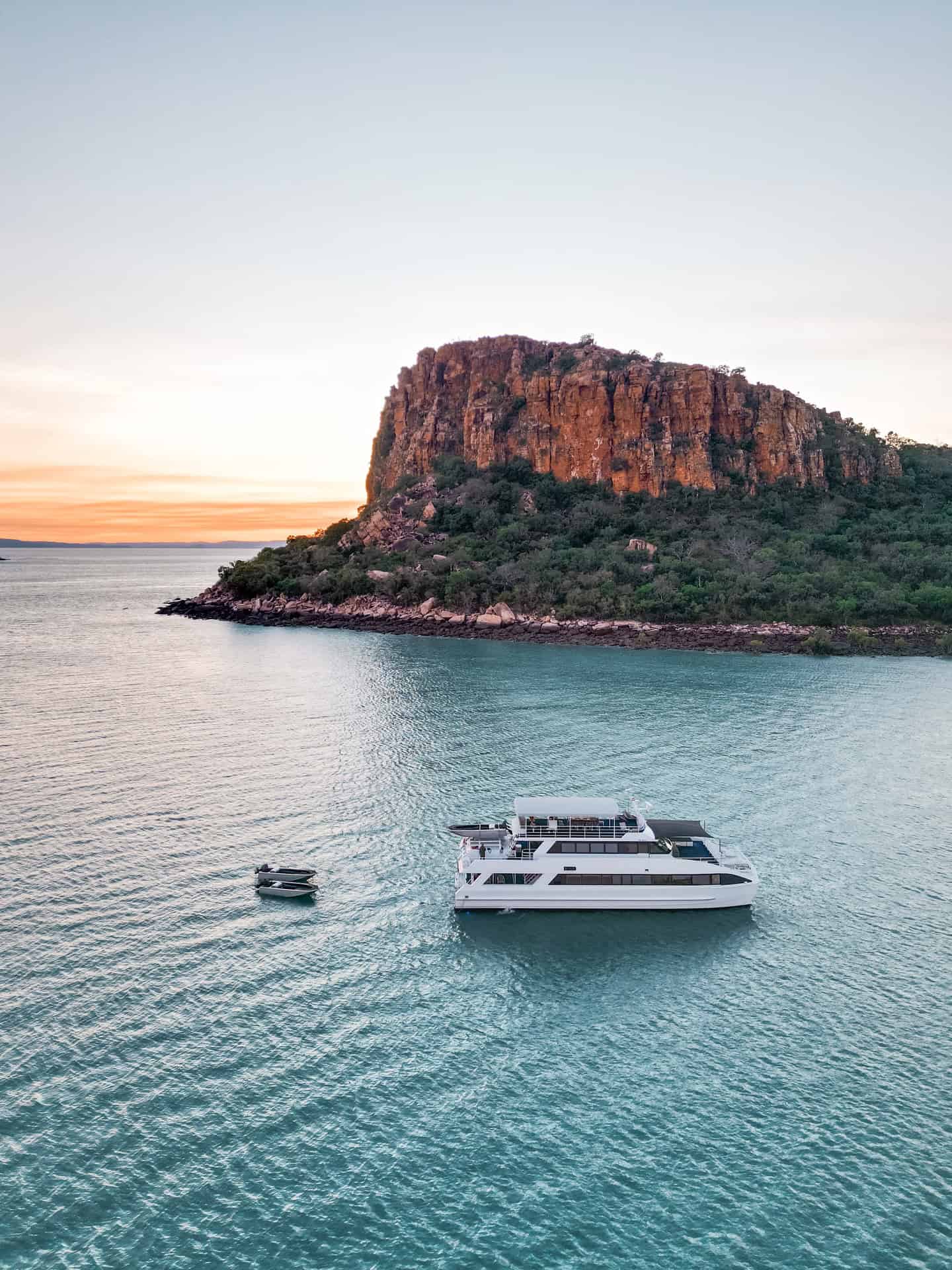 A white yacht floats near a rocky island cliff at sunset, with a small boat nearby on calm water, capturing the tranquil beauty of a Solar Eclipse Cruise.