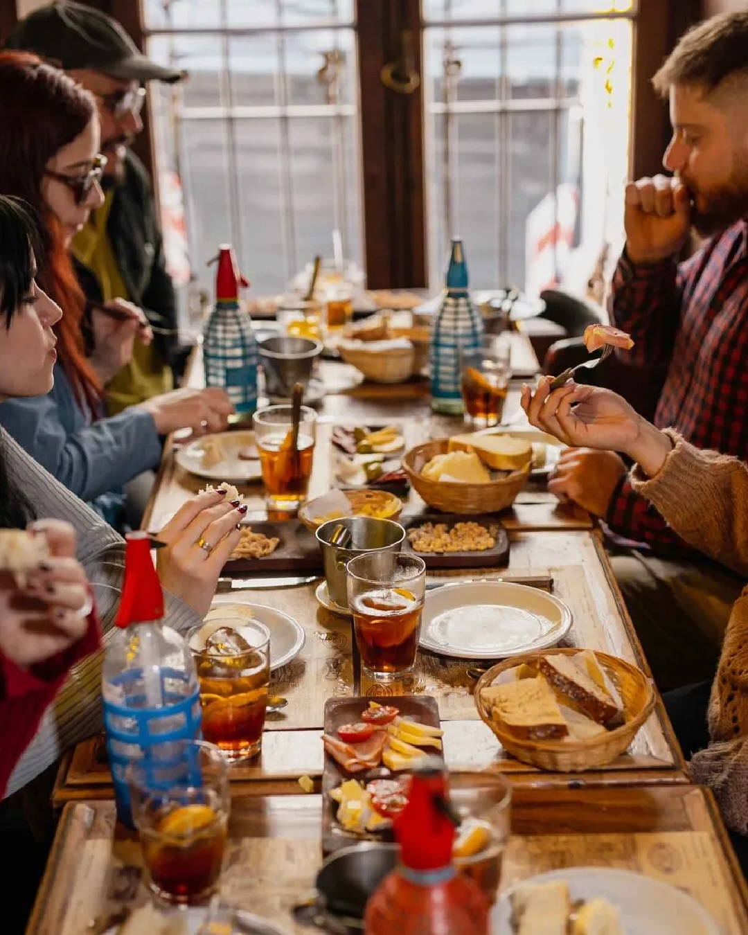 A group of people enjoy a shared meal with drinks, bread, and snacks around a rustic wooden table, evoking the lively spirit of a food tour.