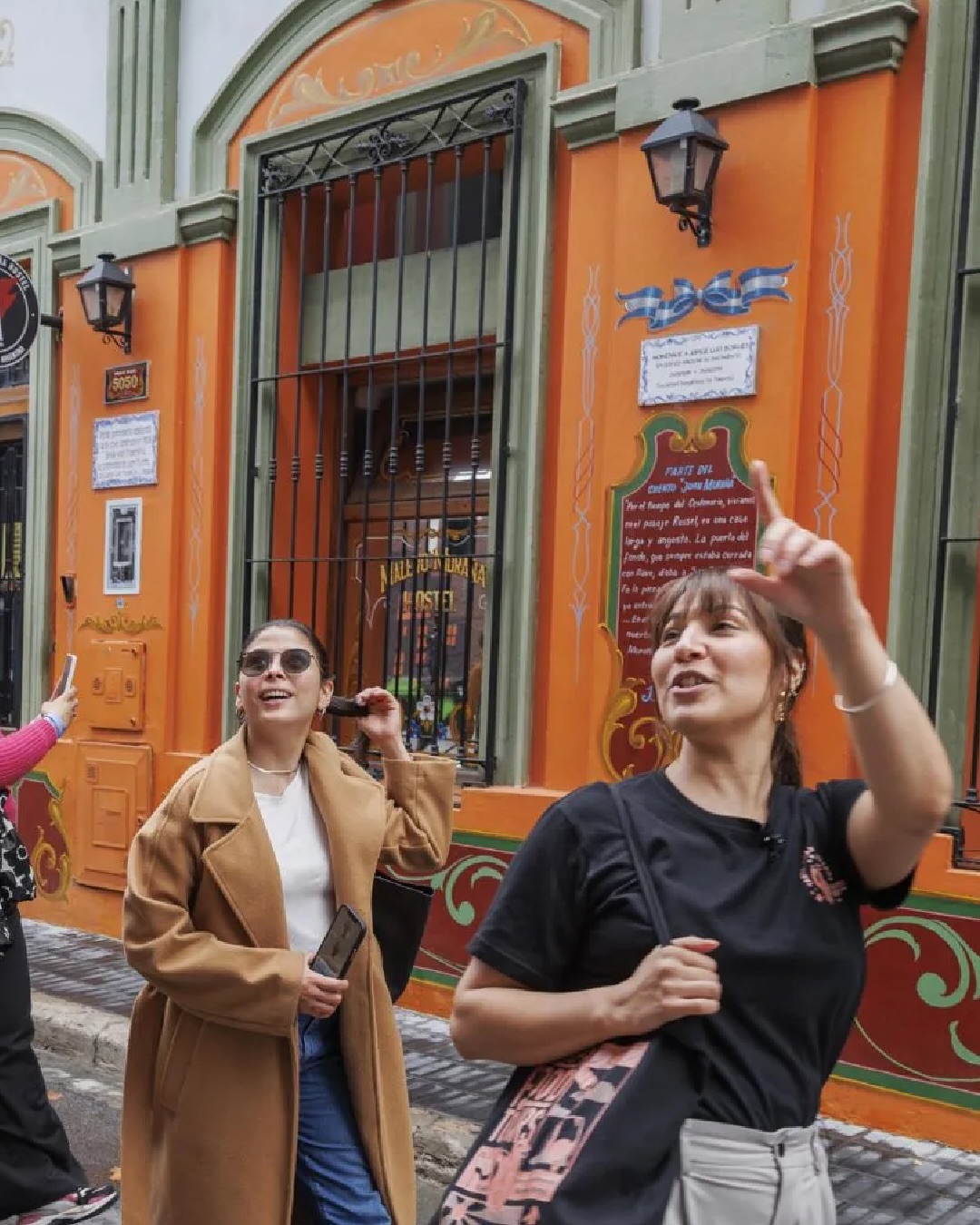 Two women smile and gesture while walking past a brightly painted orange and green building on a city street, clearly enjoying their food tour through the vibrant neighborhood.