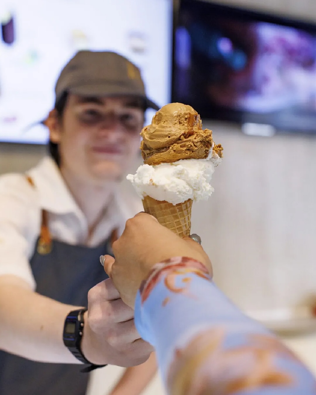 A person hands an ice cream cone with two scoops to someone at an ice cream shop counter, offering a sweet highlight on their food tour.