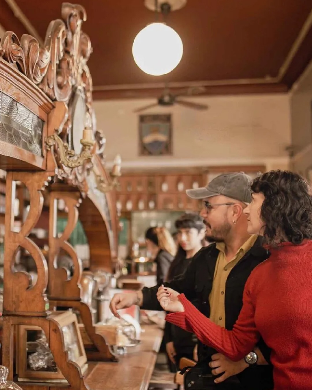 A woman in red points at an ornate wooden bar as a man in a cap looks on, capturing the lively spirit of a vintage-style cafe during their food tour.