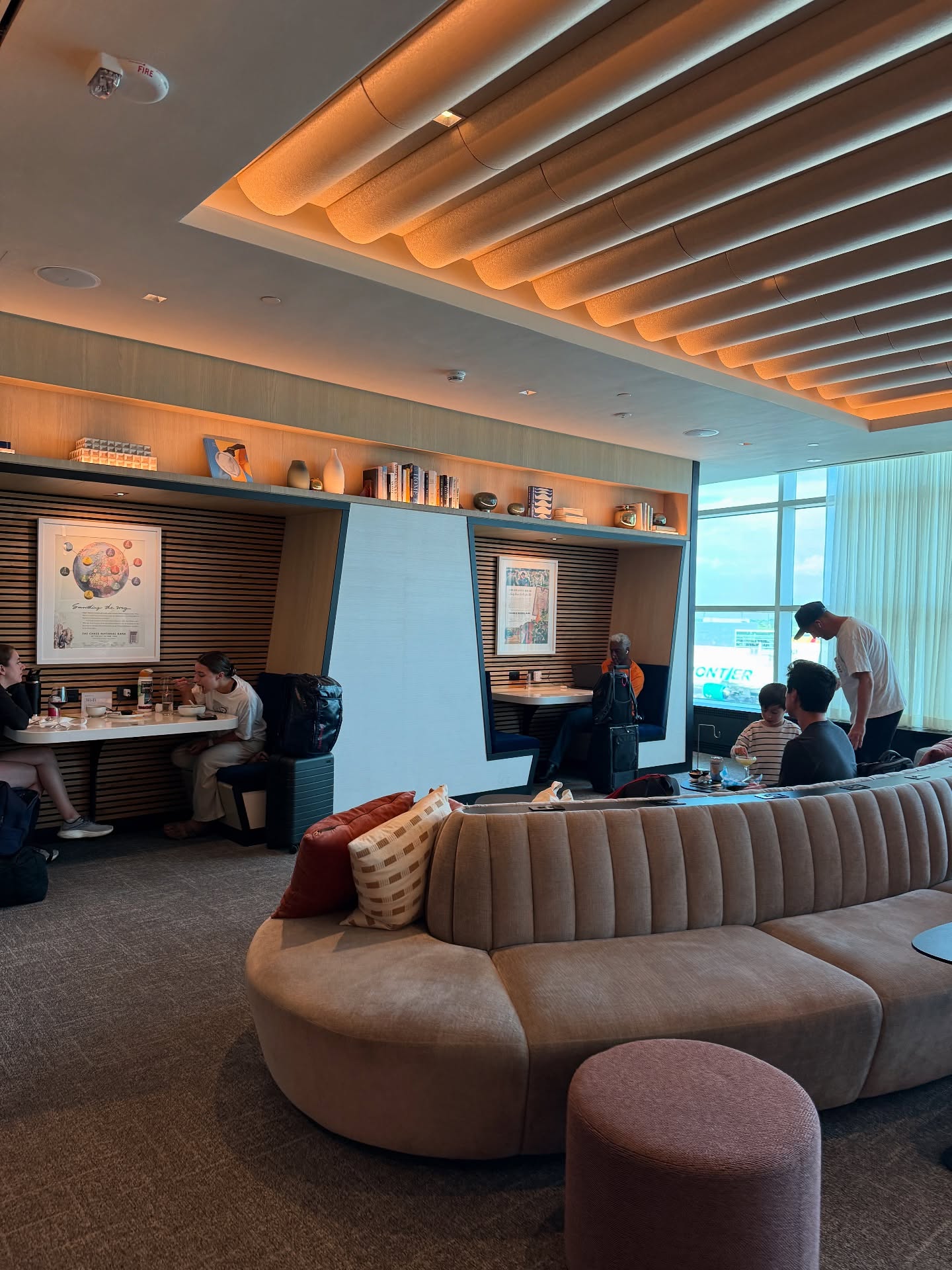 Modern airport lounge with people sitting in booths and on couches, enjoying various airport amenities, with large windows in the background.
