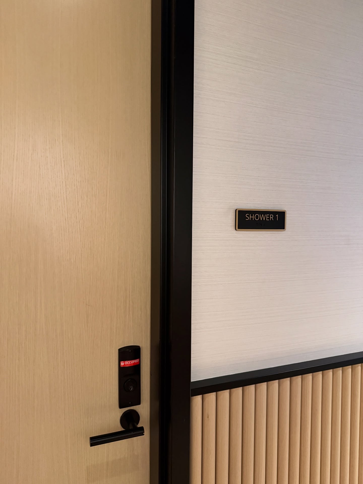 Light wood door with a keypad lock beside a sign reading “Shower 1” on a light wall with wood paneling below, offering airport amenities for travelers seeking comfort and privacy.