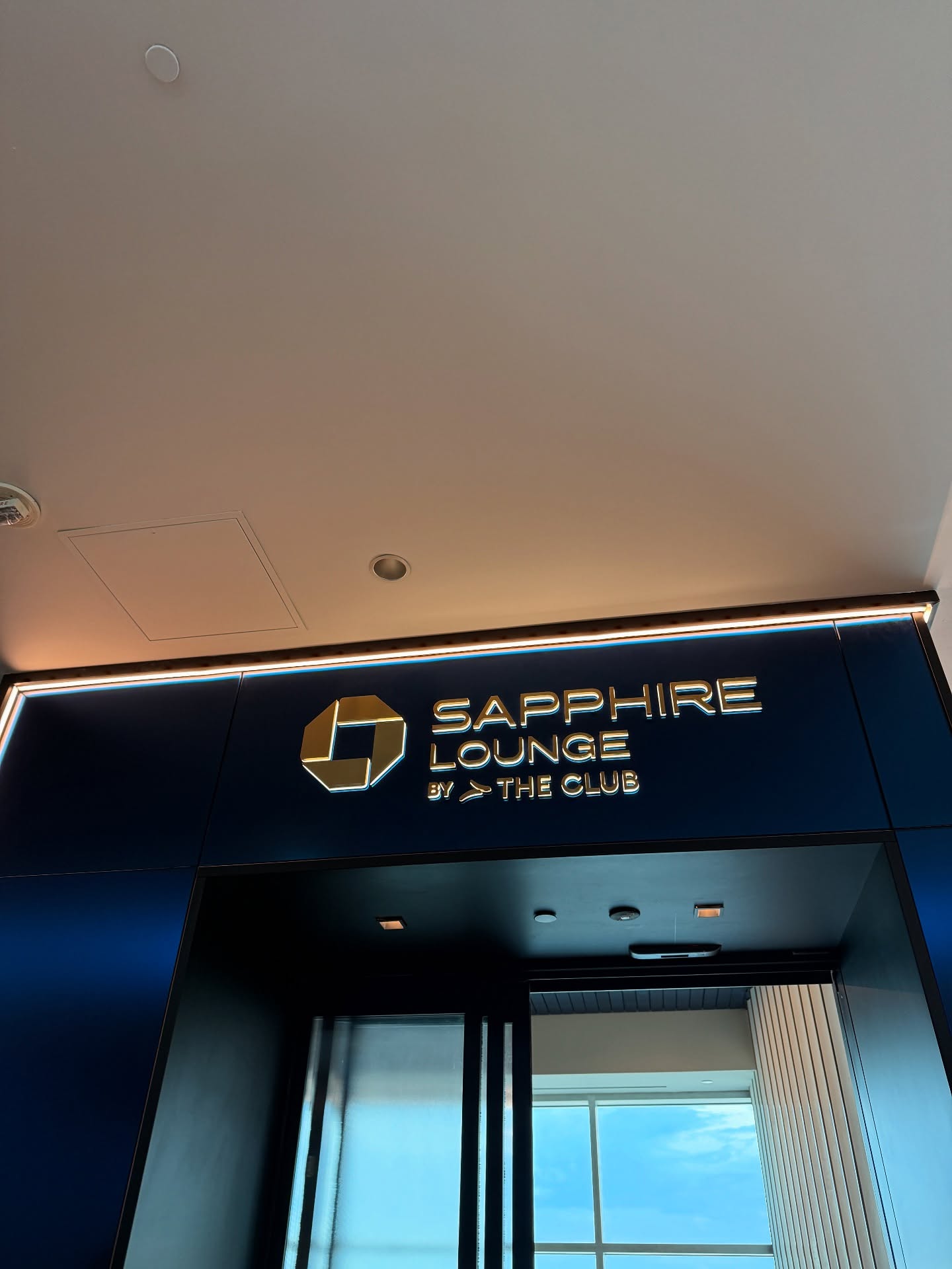 Entrance to Sapphire Lounge by The Club, featuring a gold logo and modern blue wall, showcases premium airport amenities for travelers seeking comfort and style.
