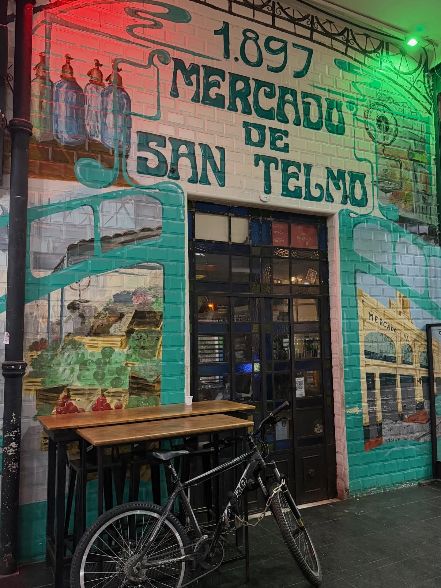 A bicycle is parked in front of a colorful mural reading “Mercado de San Telmo 1897” on a brick wall, inviting passersby to explore the vibrant market—an ideal starting point for an unforgettable food tour.