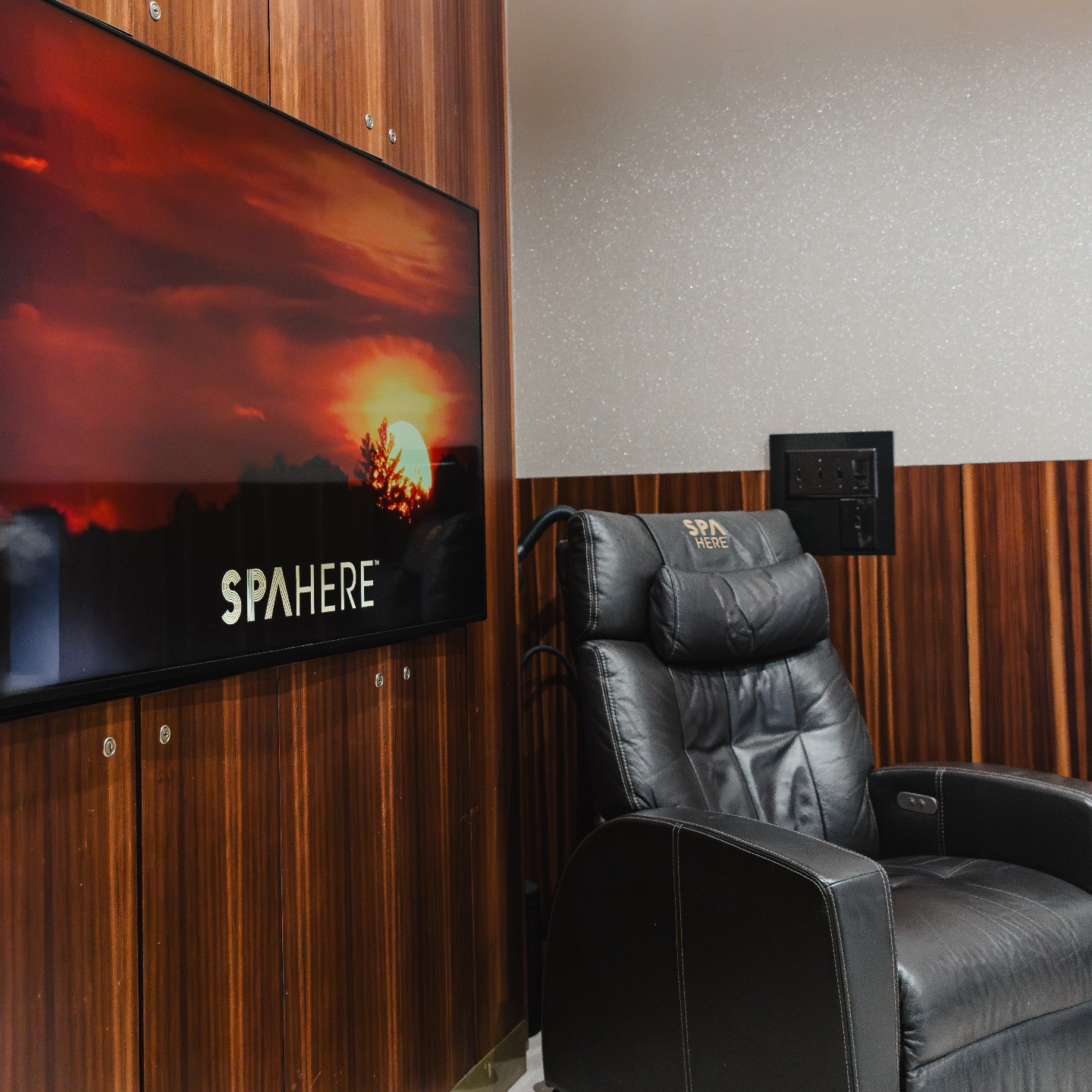 Black leather spa chair beside a wall-mounted TV displaying a sunset and the word “SPA HERE,” offering travelers relaxing airport amenities.