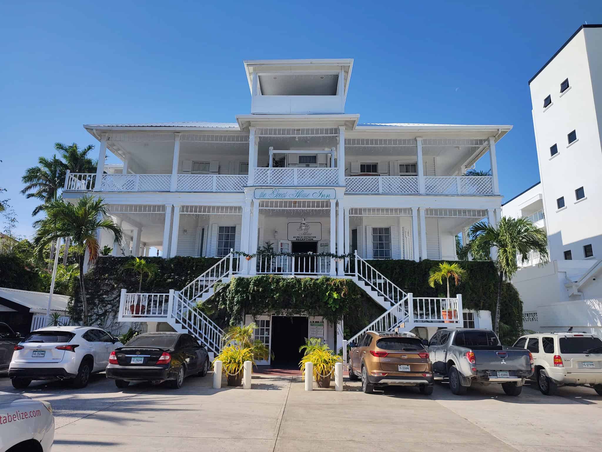 A white three-story inn with balconies, stairs, parked cars, palm trees, and clear blue sky offers a relaxing retreat after a day of exploring the Belize reef and ruins trip.