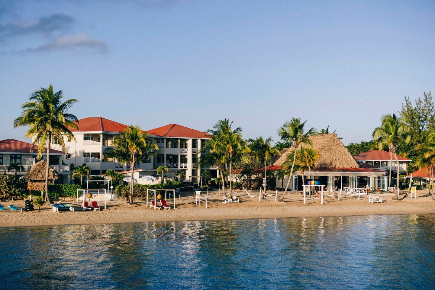 Beachfront resort with palm trees, lounge chairs, and buildings with red roofs, all by calm water.