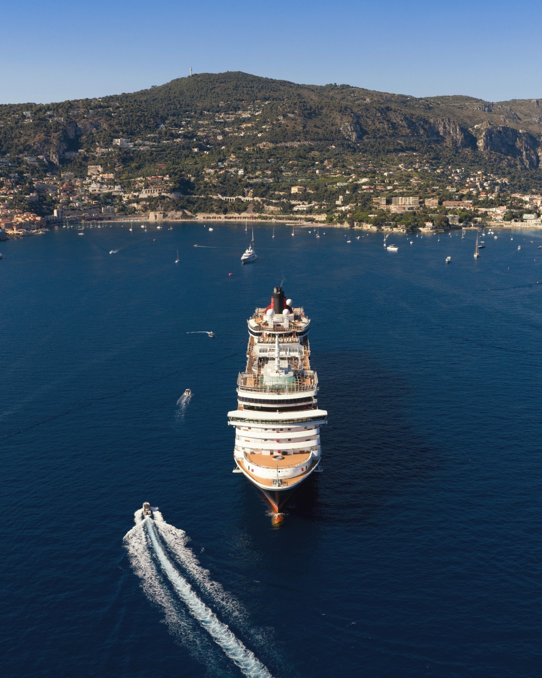 A large cruise ship sails toward a coastal town, with hills and buildings in the background and boats nearby, as part of an unforgettable Solar Eclipse Cruise experience.
