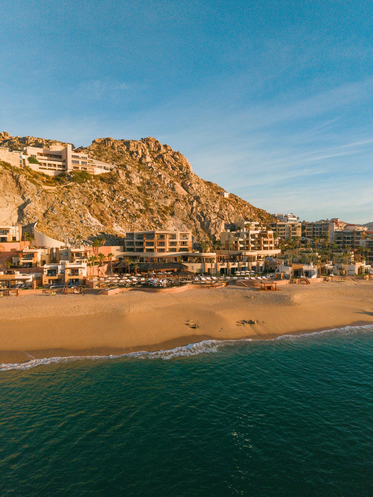 Beachfront resort buildings at the base of rocky cliffs beside calm ocean water under a clear sky, offering one of the best European destination dupes for travelers seeking idyllic seaside escapes.