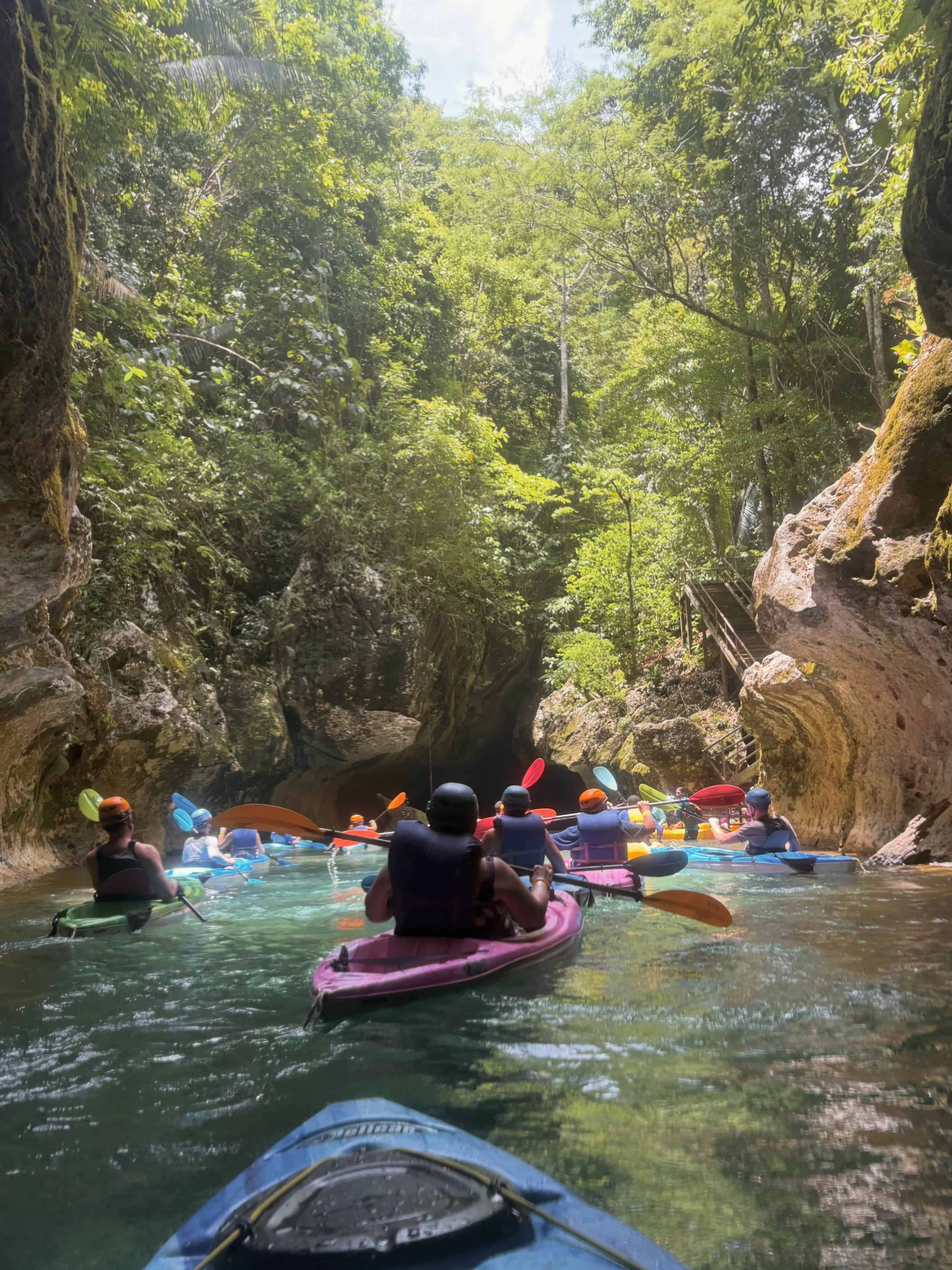 People kayaking through a lush, narrow canyon with green water and sunlight filtering through trees above, experiencing the natural wonders that make a Belize reef and ruins trip unforgettable.