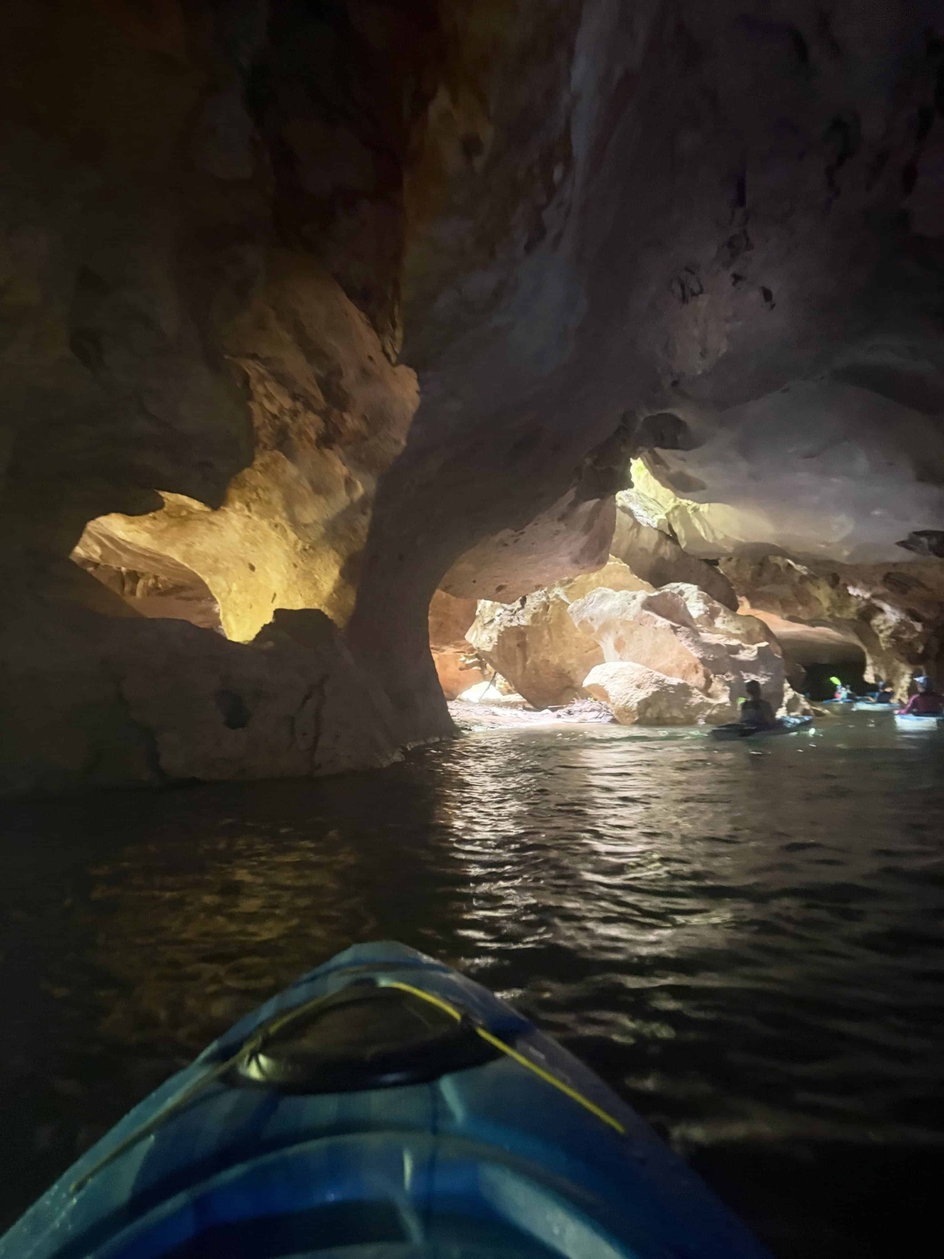 A blue kayak enters a dimly lit cave with rocky walls and water, other kayakers visible ahead on their Belize reef and ruins trip.