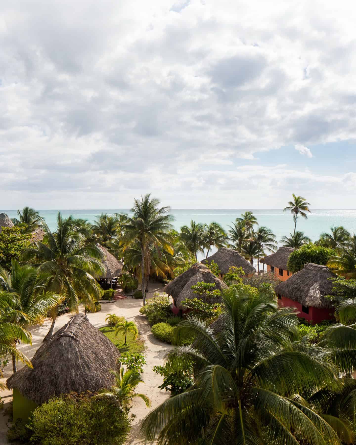Thatched-roof huts among palm trees by a turquoise ocean under a cloudy sky evoke the tranquil beauty of a Belize reef and ruins trip.