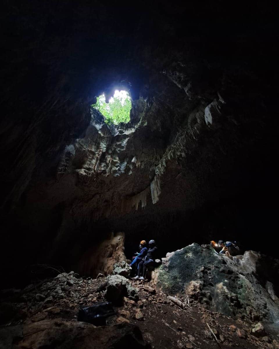 Two people sit on rocks inside a dark cave with a large opening above letting in daylight and greenery, enjoying a tranquil moment during their Belize reef and ruins trip.