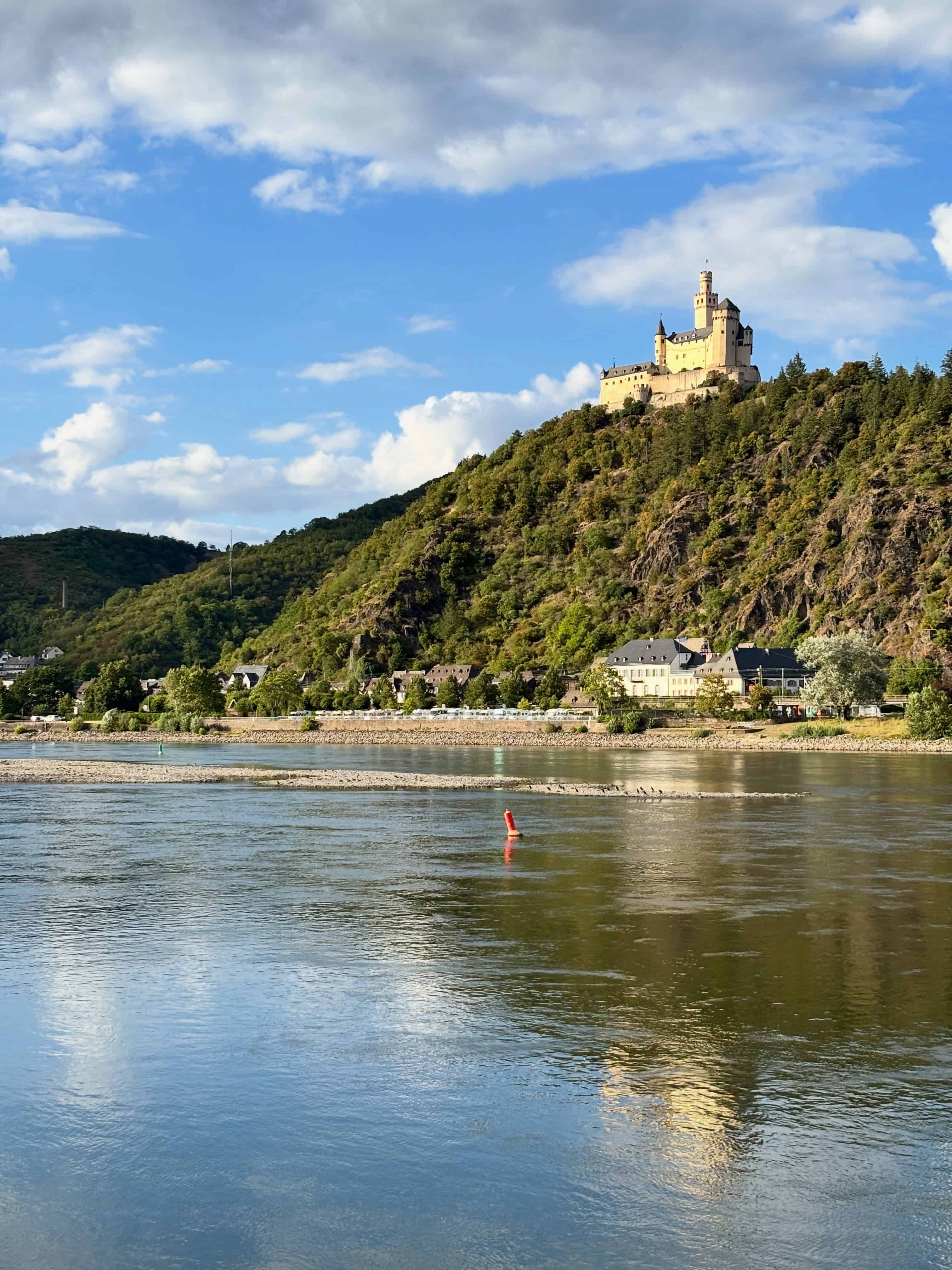 A castle sits atop a green hill, overlooking a calm river under a blue sky with scattered clouds&mdash;an idyllic setting, perfect for enjoying the wonders of nature or embarking on a Solar Eclipse Cruise along the tranquil waters below.
