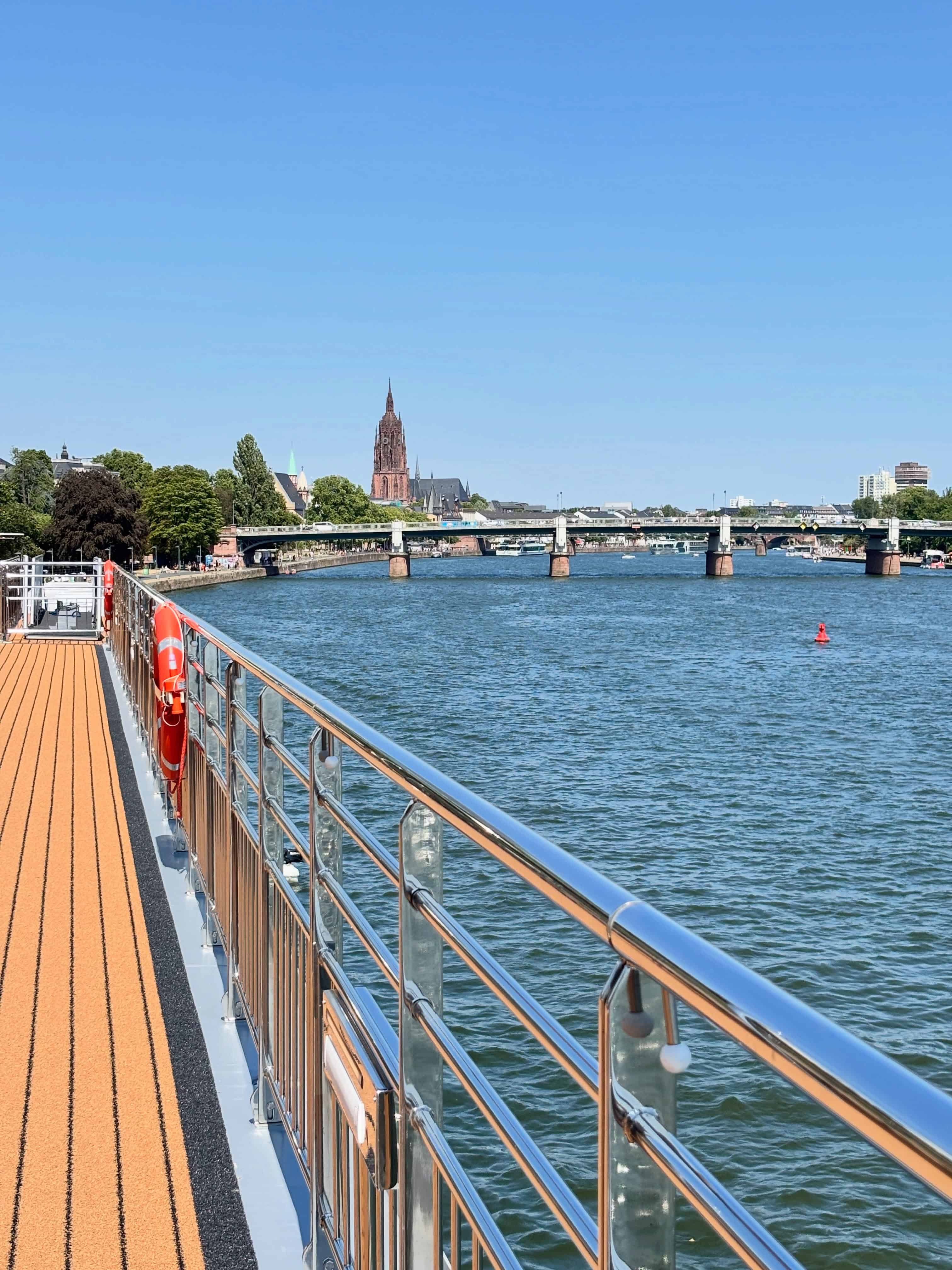 View from a boat deck on a river, with a bridge and a distant cathedral under a clear blue sky&mdash;an ideal setting for your Solar Eclipse Cruise experience.